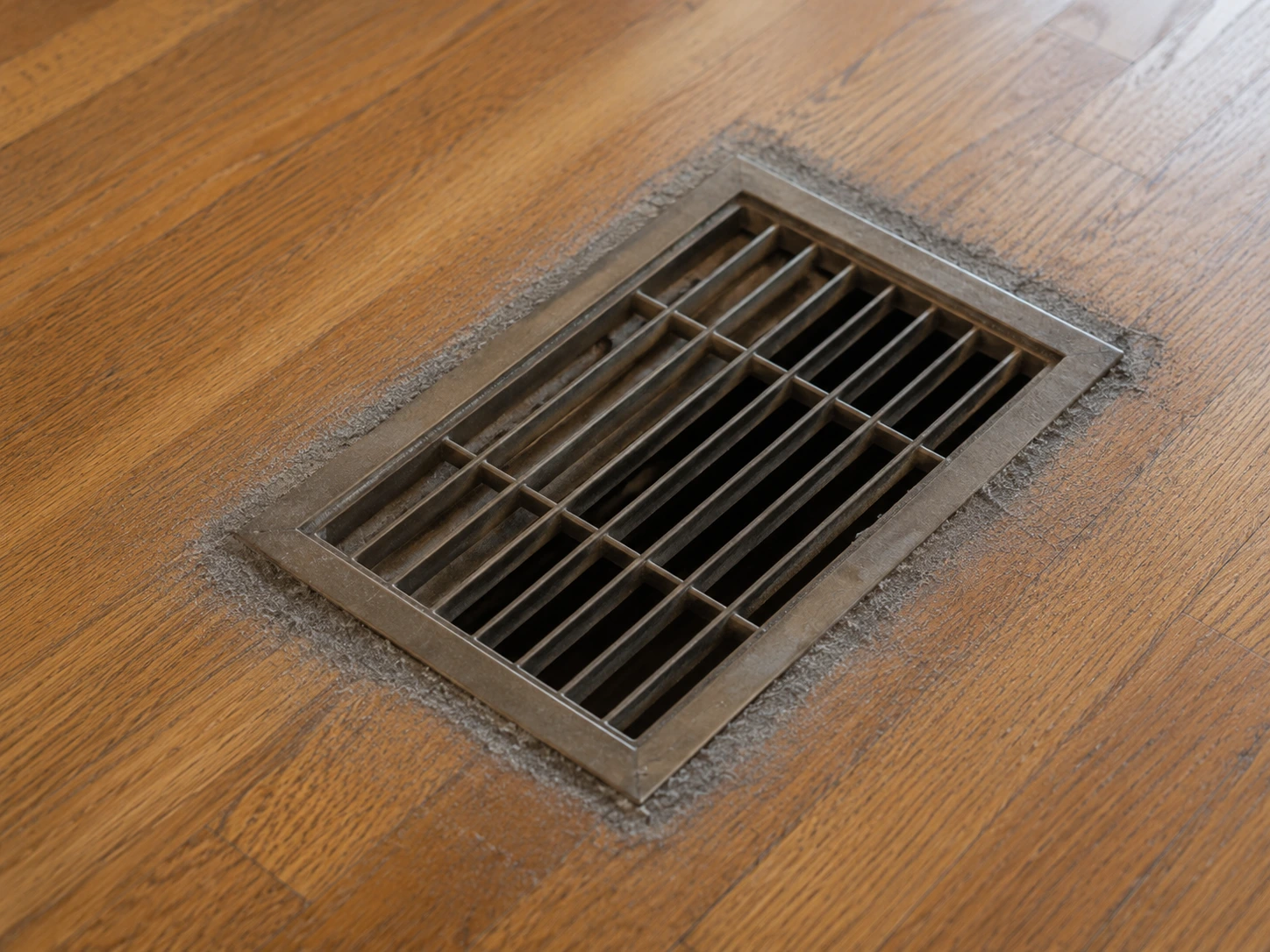 Floor air vent cover with grey dust buildup around the edges on hardwood flooring